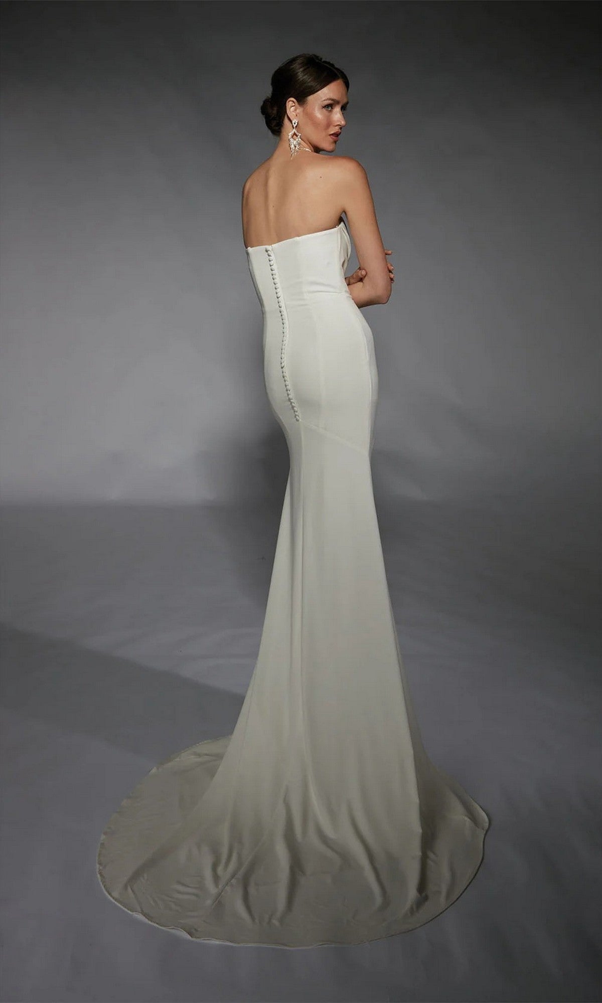A woman faces away from the camera, wearing the Alyce 7105 long white dress—a strapless formal gown with a fitted bodice and flowing train—against a plain gray background.